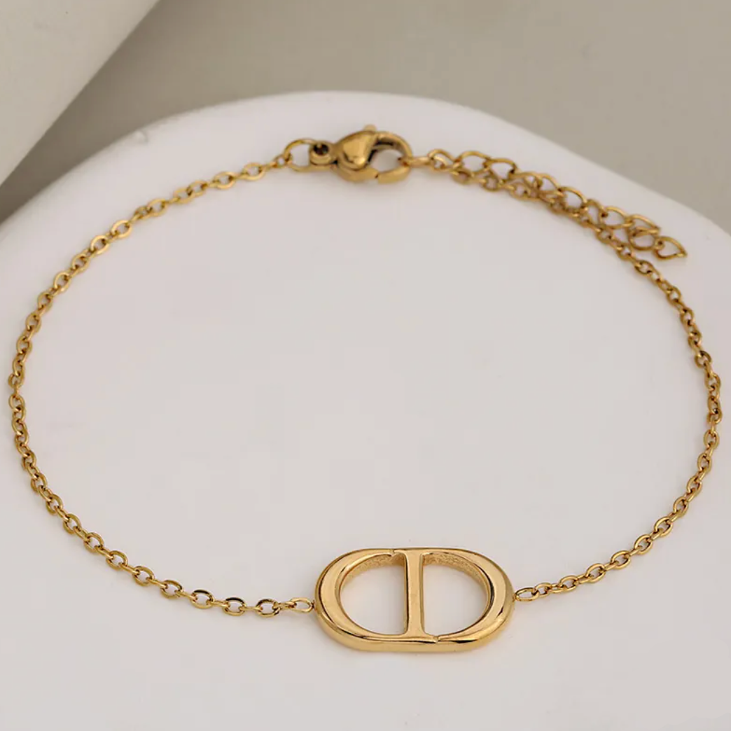 A gold-tone chain bracelet with a large interlocking D-shaped centerpiece, displayed on a white curved stand. The clasp and extender chain are visible, and text in the bottom right reads 'L:20cm+3cm'.