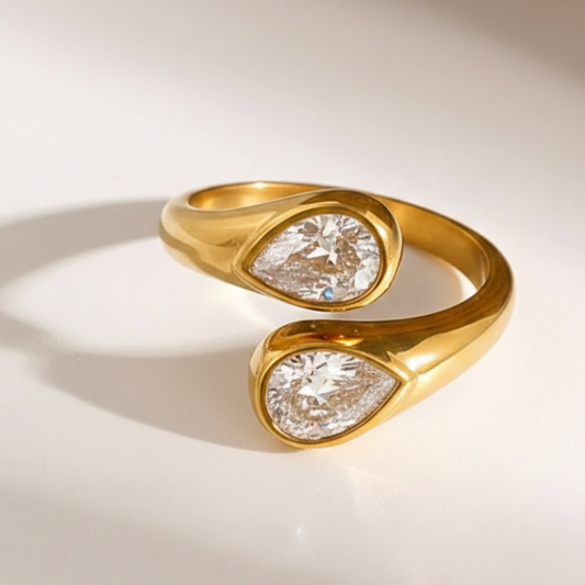 Two wrap-style rings with teardrop-shaped clear gemstones, one in silver-toned metal and one in yellow gold-toned metal, displayed on a light cream surface.